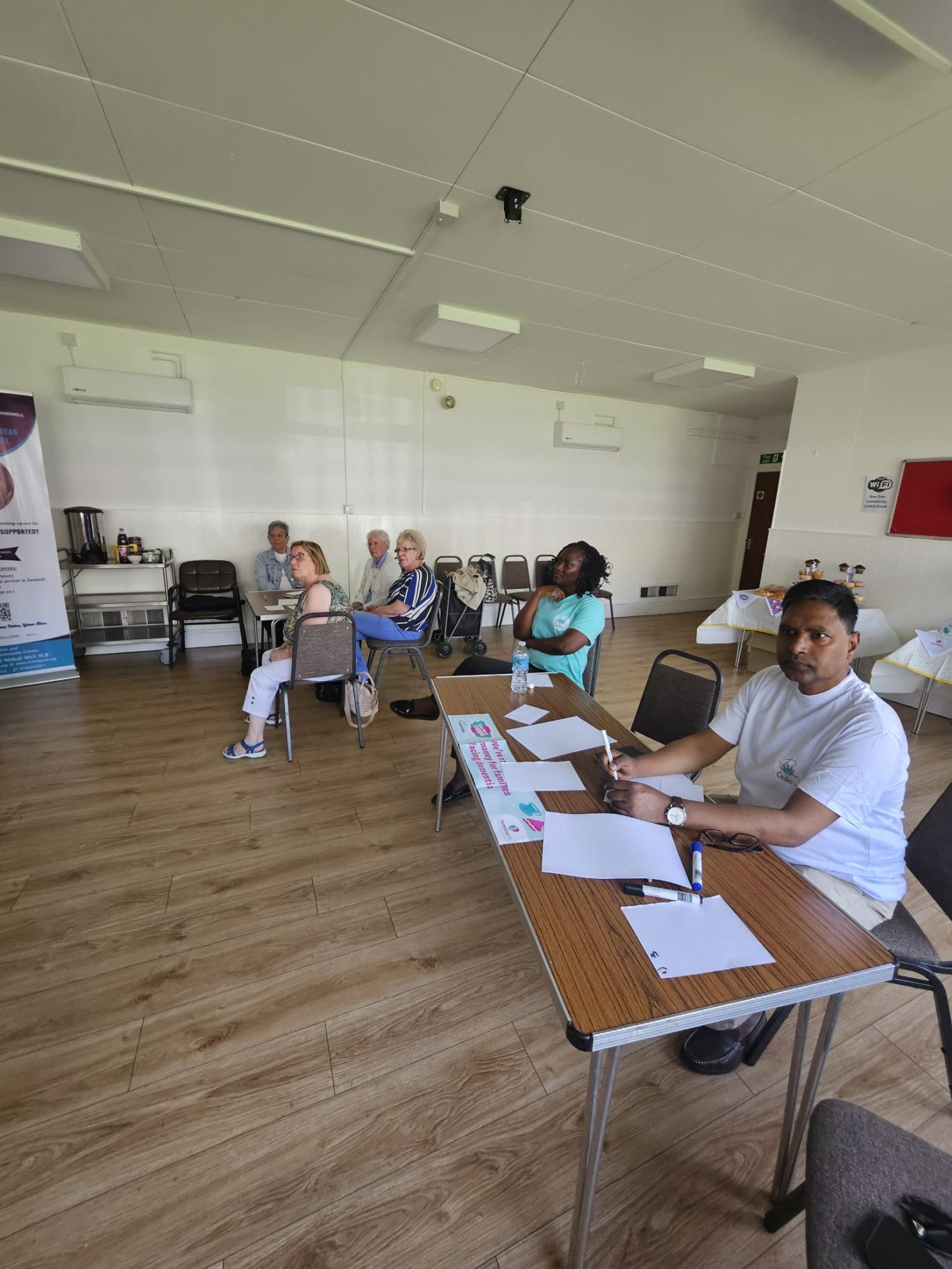 A group of people seated in a room with desks, engaged in a meeting or workshop.